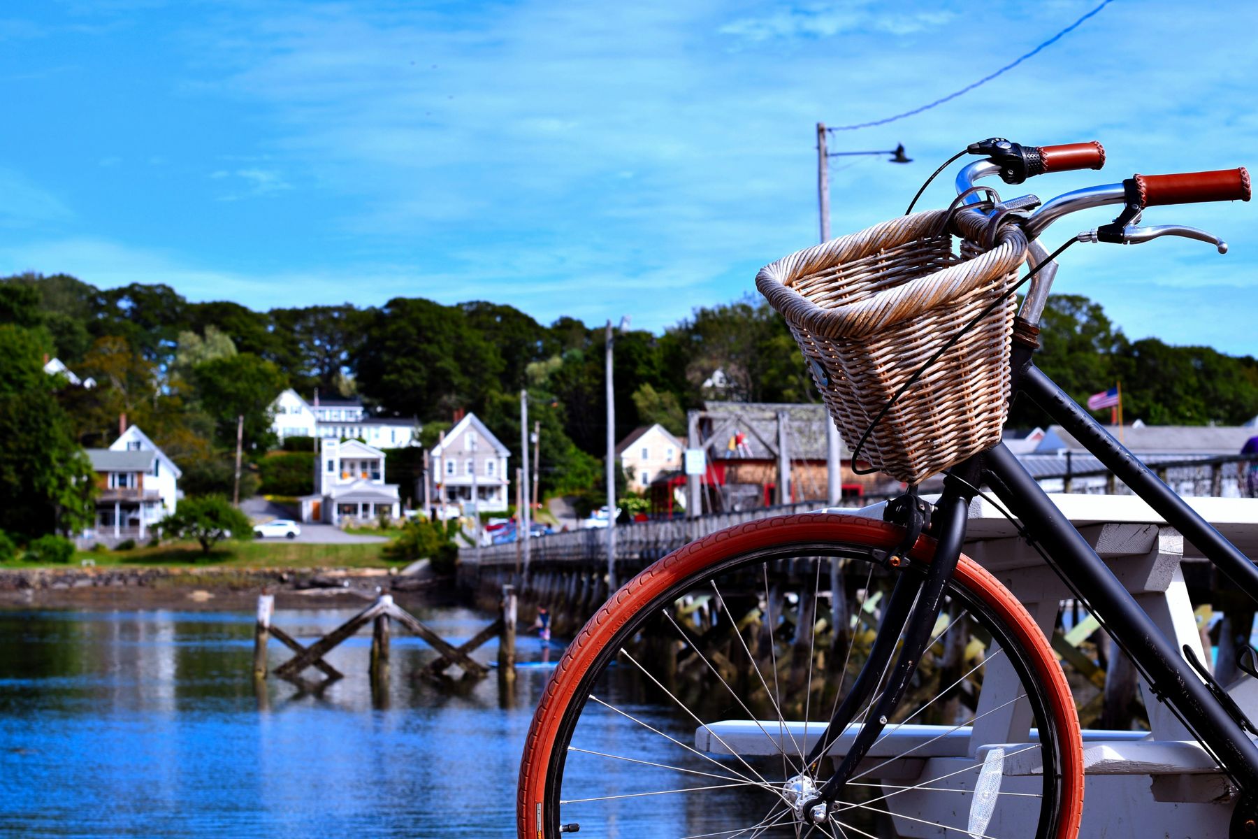 A black bicycle with a wicker basket and red tires parked on a harbor footbridge, white clapboard houses on the far shore