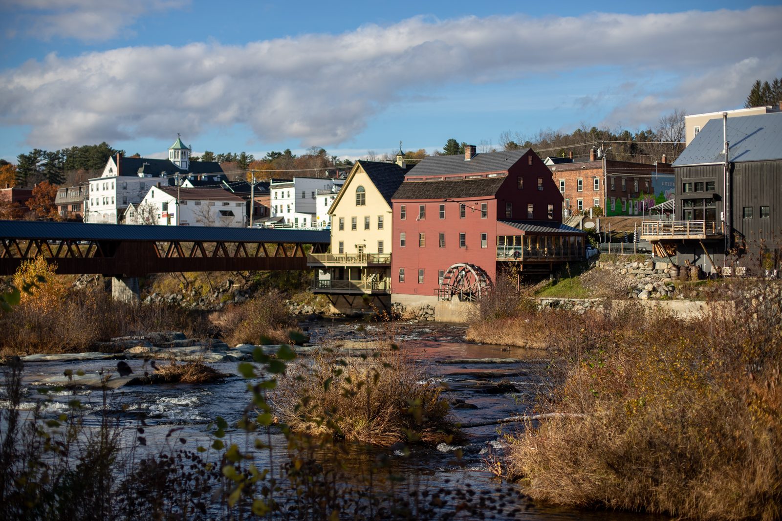 A covered bridge crossing a rocky river beside historic mill buildings in a small town